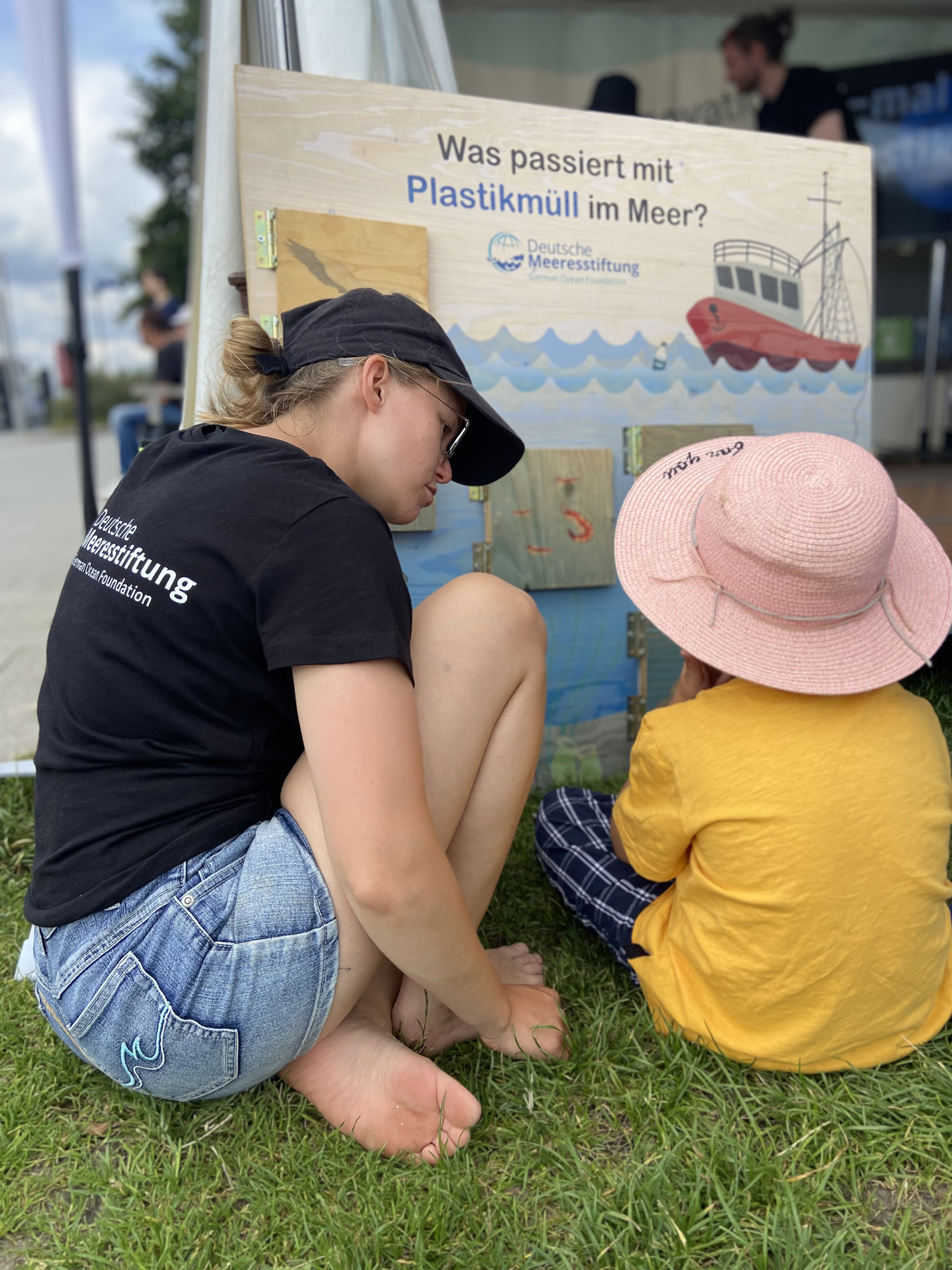 Eine Frau und ein Kind sitzen vor einem Plakat mit dem Text Was passiert mit Plastikmüll im Meer