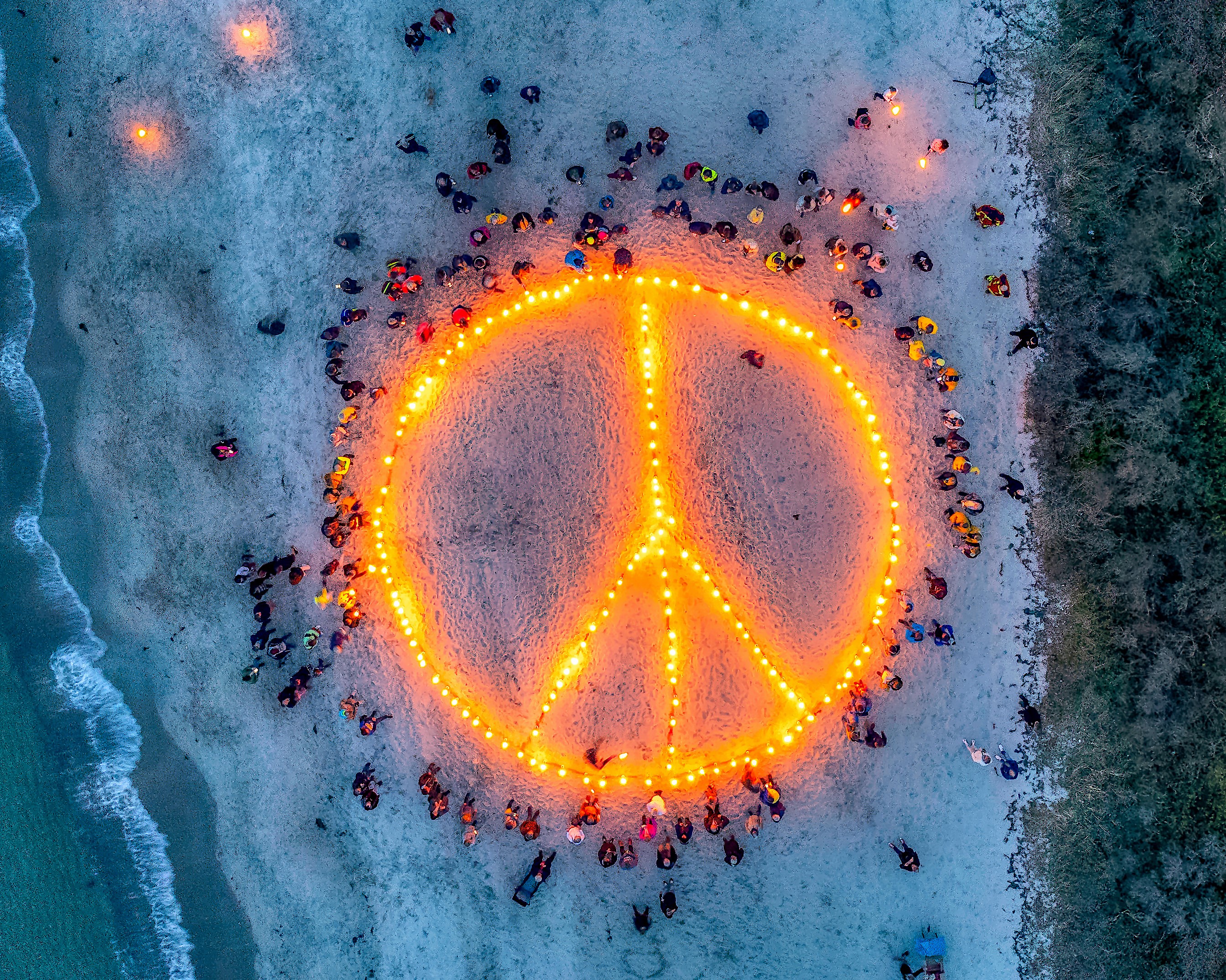 Luftaufnahme: Gigantisches Friedenssymbol aus Feuer im Sand am Strand, viele Menschen stehen darum herum