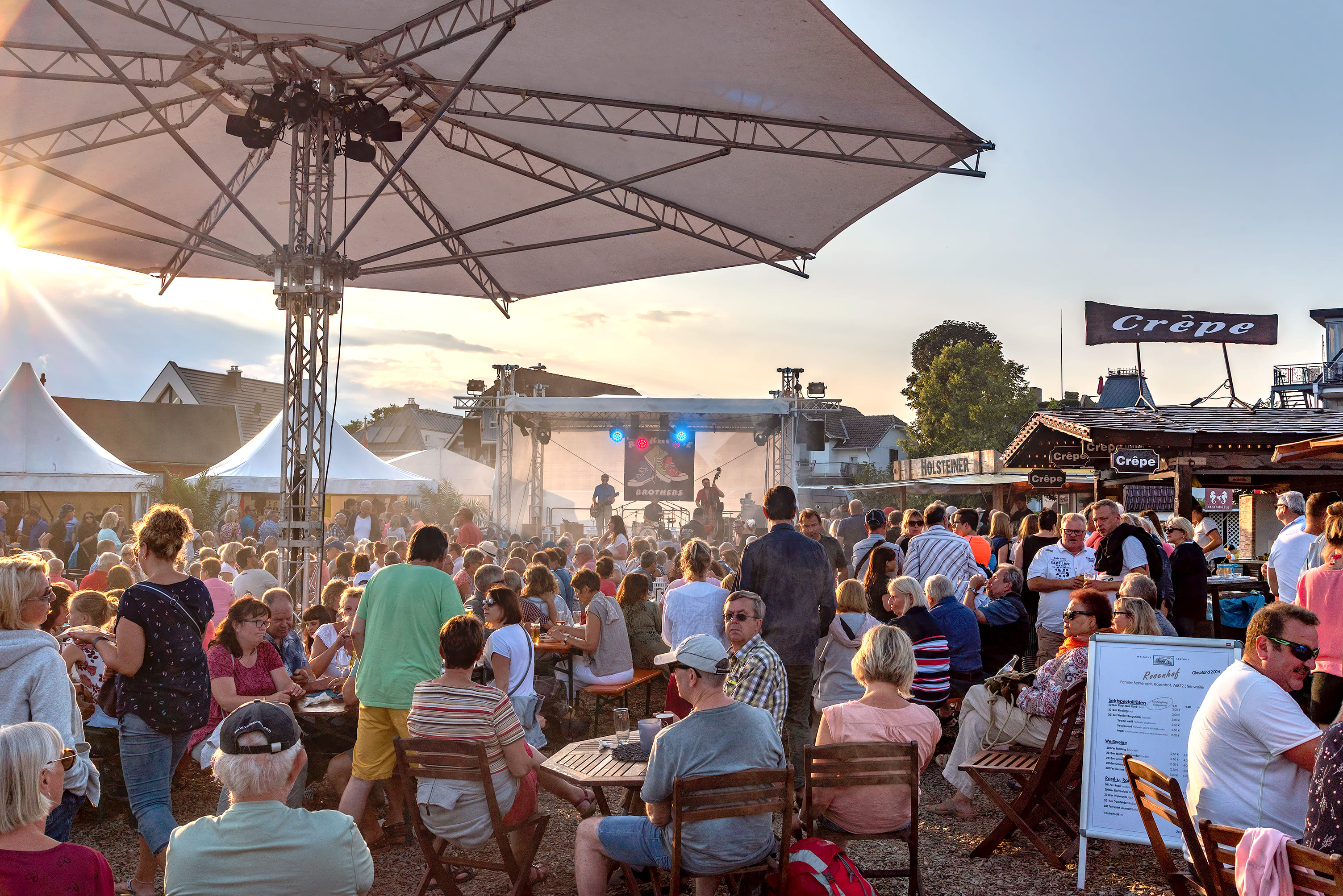 Große Veranstaltung in der Nachmittagssonne. Viele Menschen sitzen an Tischen und auf einer Bühne spielt eine Band.