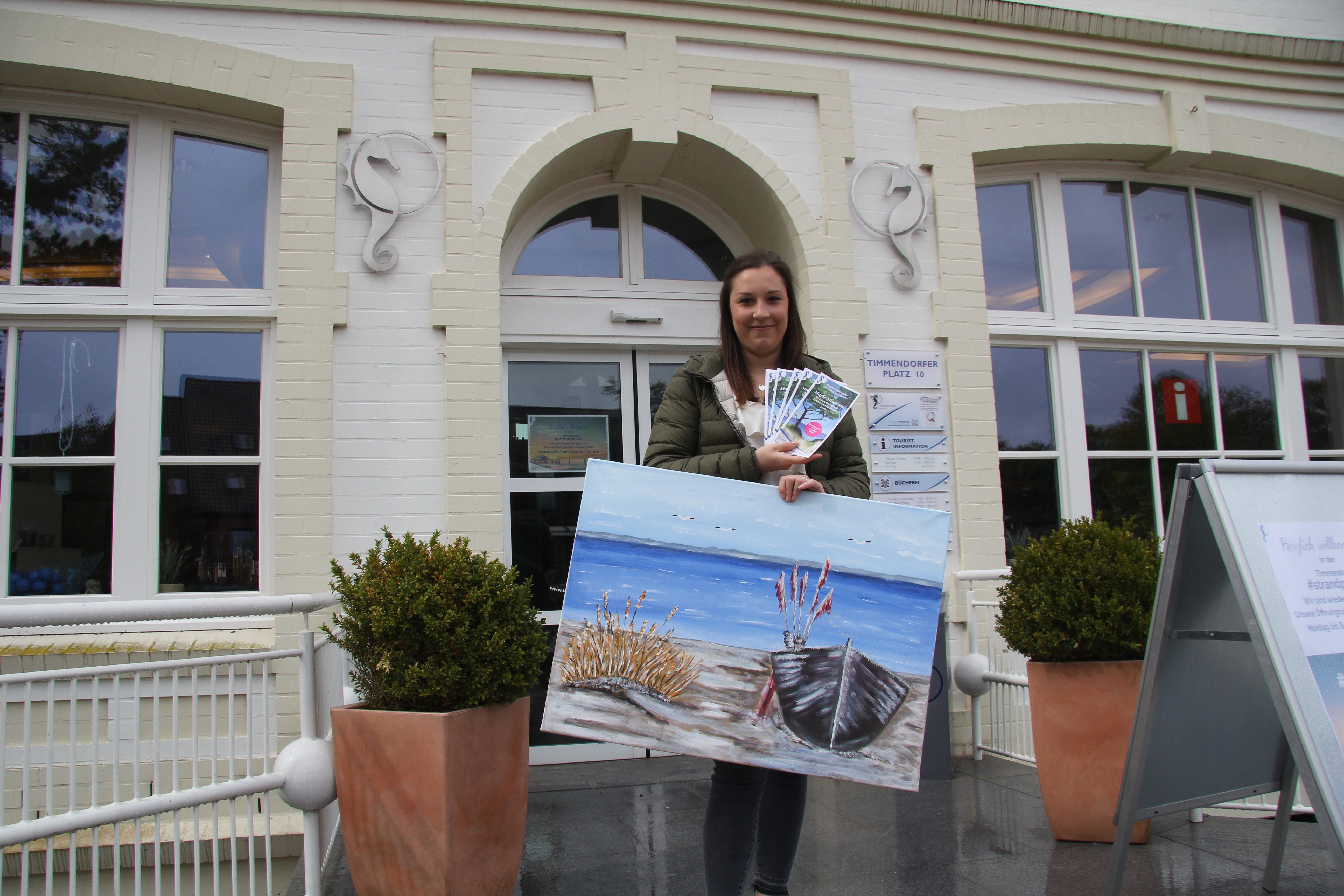 Frau steht mit einem großen Bild von einem Ruderboot und einer Hand voll Flyer draußen vor einer Eingangstür