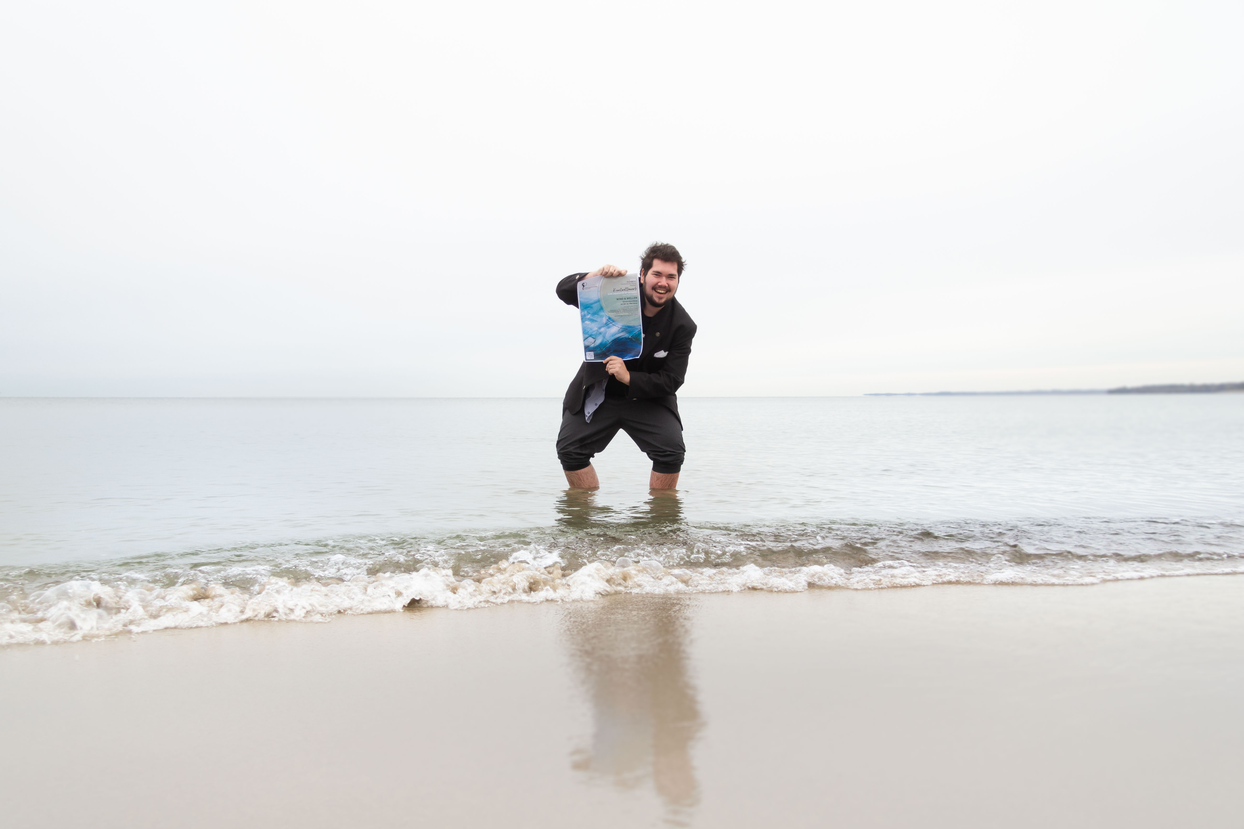 Der Auszubildende Melvin Kamp mit dem Plakat für den Kunstwettbewerb 2024 in der Hand steht mit hochgekrempelter Hose in der Ostsee